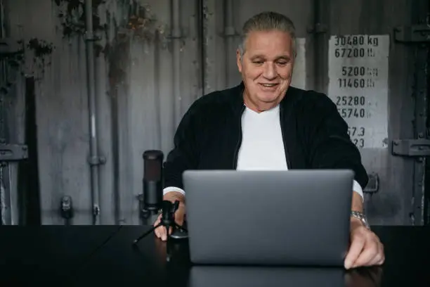 Elderly man working on a laptop with a microphone and smiling