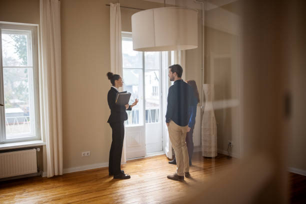 Real estate agent selling house to a young couple Full length of businesswoman discussing with customers at empty home. Female Real Estate Agent is standing with man and woman by window. They are at new apartment. house rental photos stock pictures, royalty-free photos & images