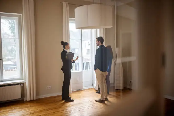 Full length of businesswoman discussing with customers at empty home. Female Real Estate Agent is standing with man and woman by window. They are at new apartment.