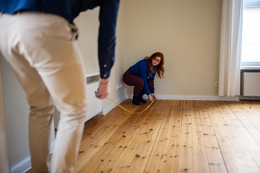 Redhead woman crouching while measuring hardwood floor with man. Couple is analyzing new home together. They are at empty apartment.