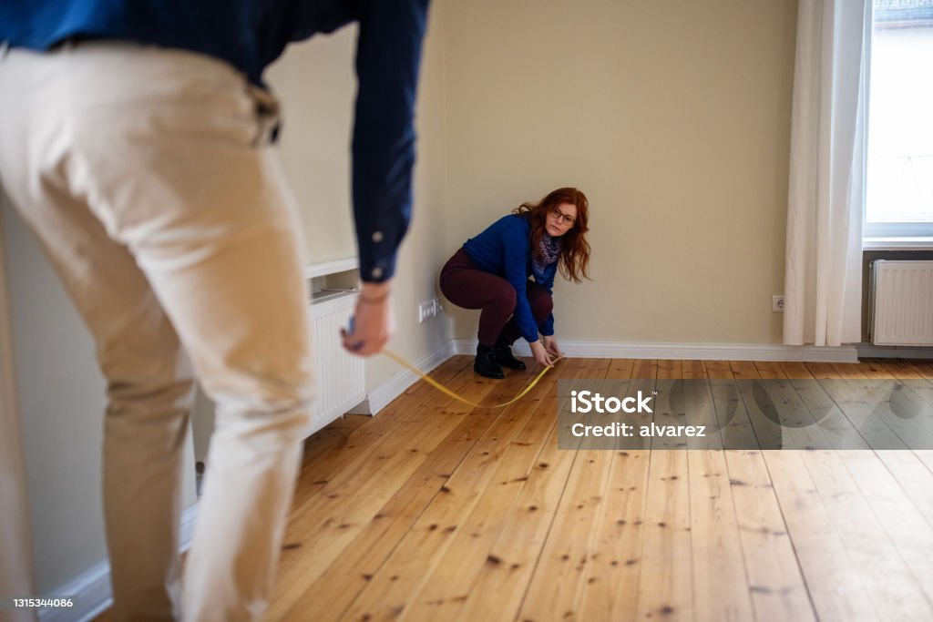 Woman crouching while measuring floor of new house with man Redhead woman crouching while measuring hardwood floor with man. Couple is analyzing new home together. They are at empty apartment. Measuring Stock Photo