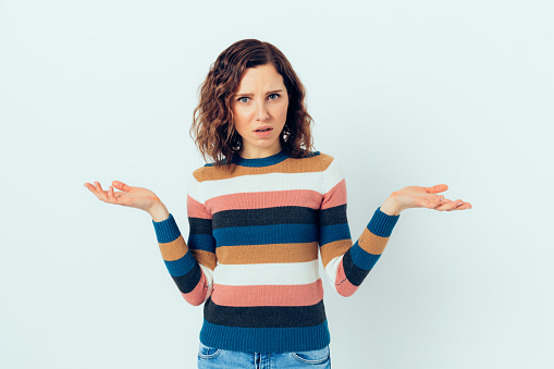 Young casual woman in disbelief shrugs shoulders with confused expression while standing against white background.