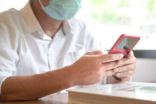 Man with face mask sitting next to windows texting on his phone. Closeup view.