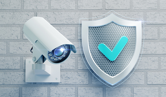External surveillance camera mounted on a brick wall beside a metallic shield, which is designed in technological style and represented as a symbol of security. 3D rendering graphics on the theme of Modern Security Technology.