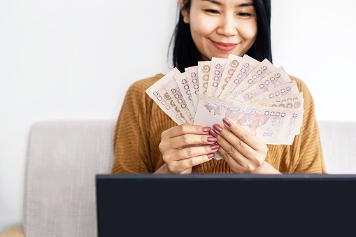 Mujer de negocios asiática mano sosteniendo billetes baht tailandeses sentados en el sofá, éxito con ganar dinero en línea photo