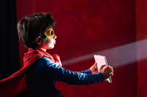 Photo of 7,5 years old schoolboy wearing super hero costume and mask and holding digital tablet against light beam. Shot indoor with a full frame mirrorless camera.