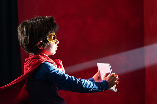 Photo of 7,5 years old schoolboy wearing super hero costume and mask and holding digital tablet against light beam. Shot indoor with a full frame mirrorless camera.