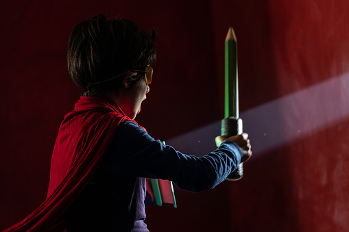 Photo of 7,5 years old schoolboy wearing super hero costume and mask and holding book and pen against light beam. Shot indoor with a full frame mirrorless camera.