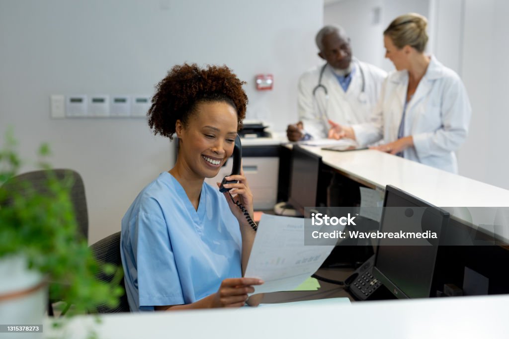 Mixed race female doctor at desk in hospital reception talking on phone and holding document Mixed race female doctor at desk in hospital reception talking on phone and holding document. medicine, health and healthcare services. Receptionist Stock Photo