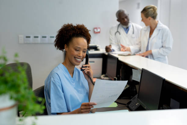 Mixed race female doctor at desk in hospital reception talking on phone and holding document Mixed race female doctor at desk in hospital reception talking on phone and holding document. medicine, health and healthcare services. receptionist stock pictures, royalty-free photos & images