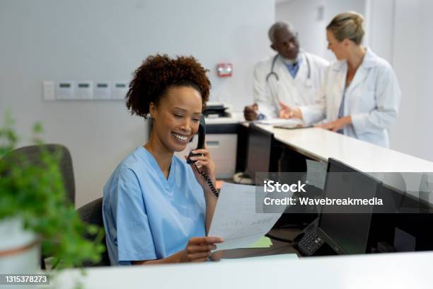 Mixed Race Female Doctor At Desk In Hospital Reception Talking On Phone And Holding Document Stock Photo - Download Image Now