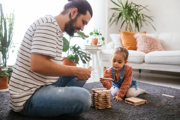Caring young dad and cute daughter playing with wooden blocks sit on floor, father enjoying teaching helping little child holding toys having fun at home. Wooden bars for a board game in a tower puzzle Jenga. Happy time