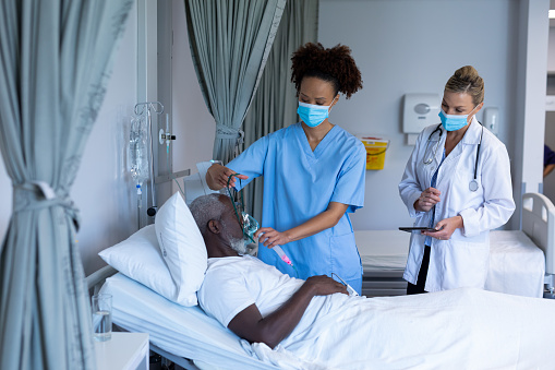 Mixed race female doctor wearing face mask putting oxygen mask ventilator on male patient. medicine, health and healthcare services during coronavirus covid 19 pandemic.