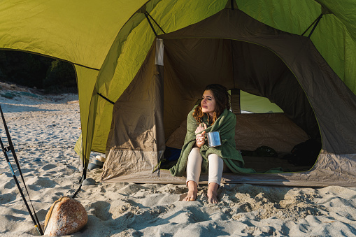 Portrait of young smiling woman inside a tent holding a mug and wearing a blanket in the morning just waked up looking at distance. Camping on the beach.