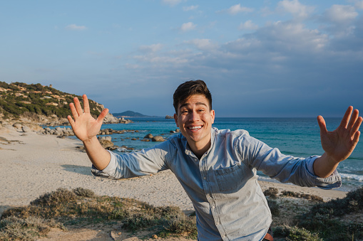 Portrait of happy joyful young latin man smiling looking at the camera with open arms with beautiful beach on background. Travel destination in Villasimius, Sardinia, Italy.