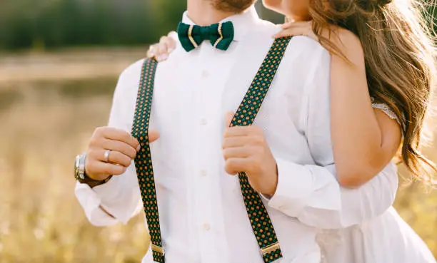 Photo of Half portrait of bride hugging groom from behind. Bride in a beautiful white dress. Groom in white shirt, green bow-tie and colorful suspenders