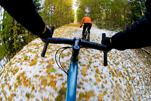 The handlebar view of two men going for a gravel bike ride on a rail grade trail in October. Gravel bikes are similar to cyclo-cross bikes with tires suited for riding on rough terrain. Some snow is visible on the ground along with the fallen aspen leaves.