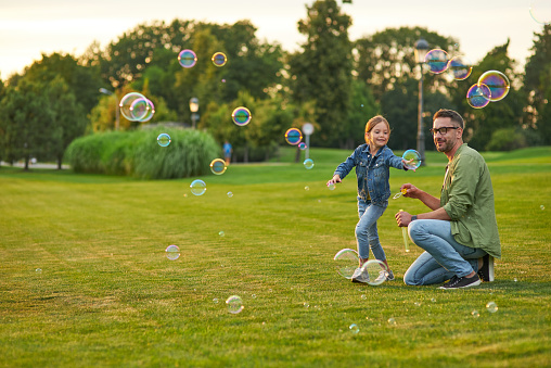 Full length shot of young father and his cute little girl having fun while blowing soap bubbles in the park, daughter and dad spending time together outdoors. Happy family and childhood concept