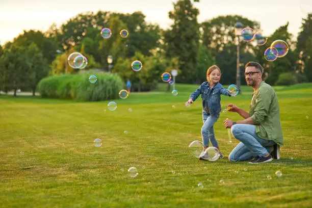 Photo of Full length shot of young father and his cute little girl having fun while blowing soap bubbles on a summer day, daughter and dad spending time together outdoors