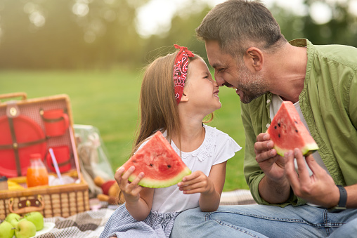 Adorable little girl playing with her loving daddy while eating watermelon, family having a picnic in the green park on a summer day. Parenthood, happy childhood concept