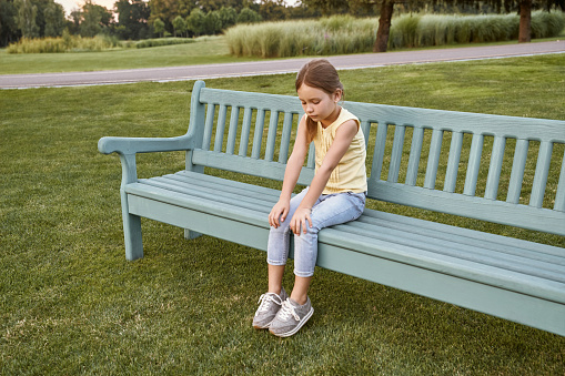 Sad little girl sitting on the bench in park on a warm day, waiting for parents outdoors. Childhood concept