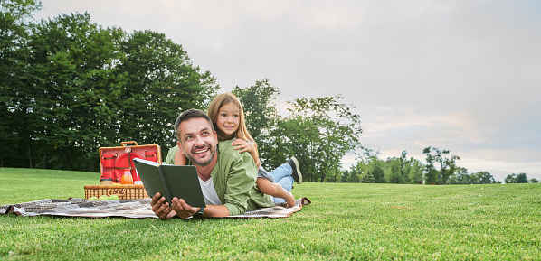 Cheerful cute little girl spending time with her father, reading a book while having picnic in the park. High quality illustration. Happy family, childhood concept