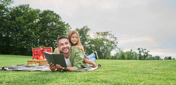 wesoła urocza dziewczynka spędza czas z ojcem, czytając książkę podczas pikniku w parku - men reading outdoors book zdjęcia i obrazy z banku zdjęć