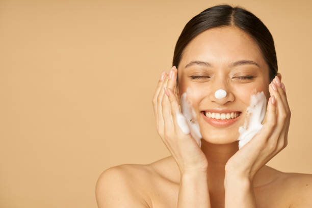 Studio portrait of pleased young woman smiling with eyes closed while applying gentle foam facial cleanser isolated over beige background Studio portrait of pleased young woman smiling with eyes closed while applying gentle foam facial cleanser isolated over beige background. Beauty products and skin care concept. Front view facial cleanser stock pictures, royalty-free photos & images
