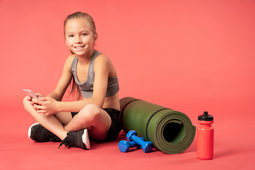 Cute female child holding smartphone and smiling while sitting near yoga mat, dumbbells and bottle of water. Isolated on red background