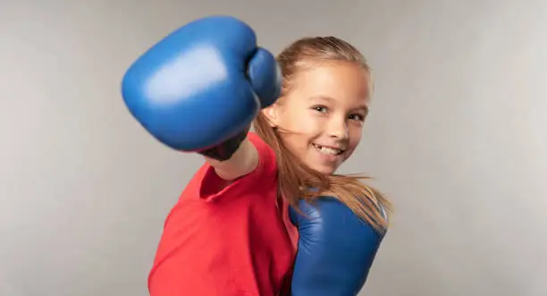 Cheerful female child in boxing gloves punching air and smiling while standing against gray background