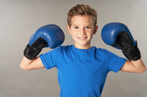 Adorable male kid boxer wearing sports boxing gloves and blue shirt while looking at camera and smiling