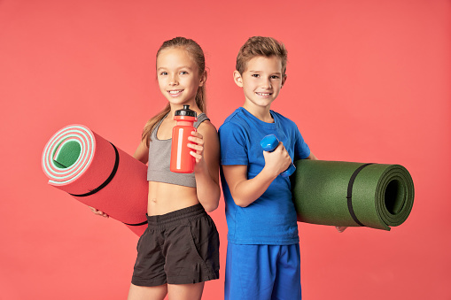 Cheerful cute girl with bottle of water looking at camera and smiling while boy holding dumbbell and yoga mat