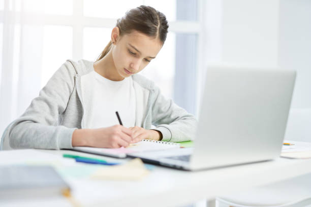 istock Concentrated latin teenage girl making notes in her notepad, using laptop while having online lesson at home 1315445161