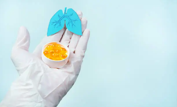 Paper lungs and white pills on doctorâs gloved hand, blue background. Omega-3 supplements help fight coronavirus and lung disease