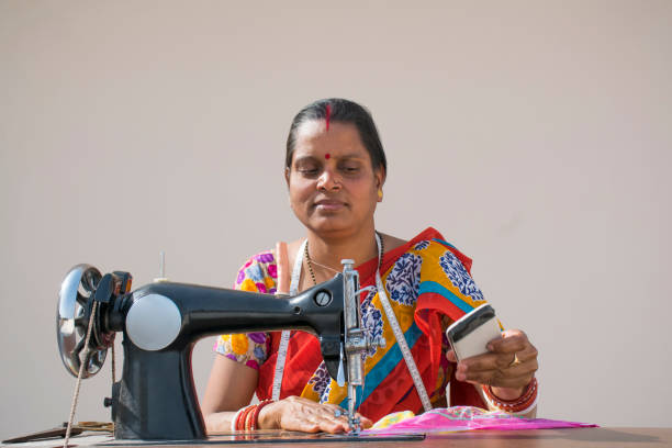 istock Indian woman stitching cloths by machine and using mobile 1315446770