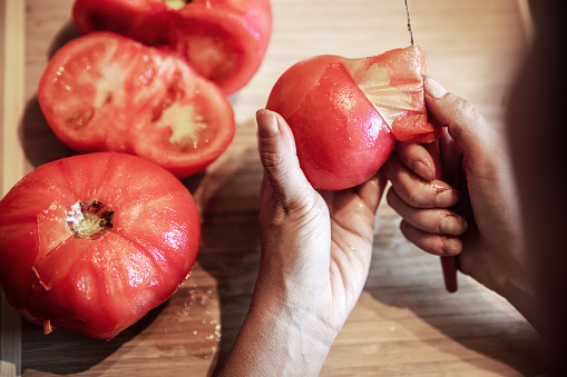 a woman is peeling a lot of red fresh and boiled tomatos with a knife, preparing for cooking a sauce