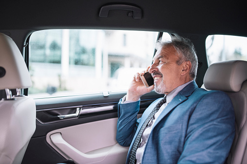 Side view of a senior entrepeneur usig his phone and smiling during business travel by a car.