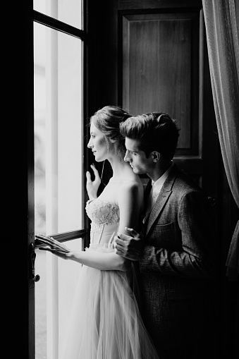 Groom hugs bride from behind at the large window of an old villa. Lake Como. Black and white photo. High quality photo