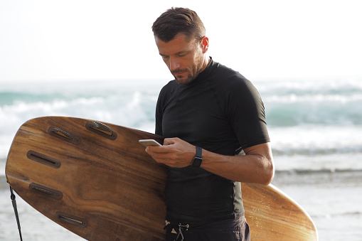A male surfer holds a brown surfboard and uses a smartphone on the beach.