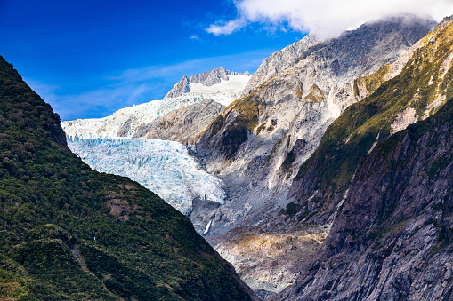 This April 2021 daylight image shows Te Tai o Wawe / Franz Josef Glacier in Westland Tai Poutini National Park, Aotearoa New Zealand. Due to a warming planet, the glacier has retreated over the decades exposing the rocky valley below. Shadows are cast by low clouds floating through the valley.