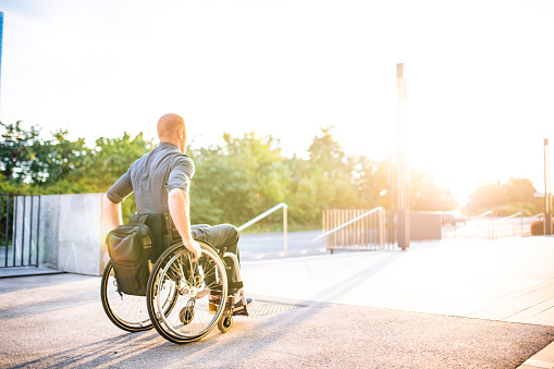 Lens flare view of Caucasian male business professional in mid 20s moving along sun-filled pedestrian pathway in wheelchair.