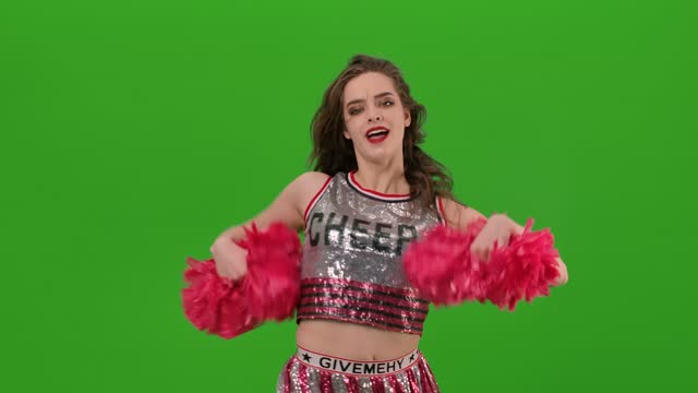 An attractive young cheerleader in uniform performs a jubilant dance with red pompoms in the studio on a green screen. Close up