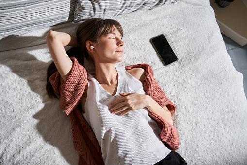 Enjoying. High angle view of the caucasian woman wearing earphones laying at the bed and listening music with closed eyes while relaxing at the great sunny morning. Stock photo