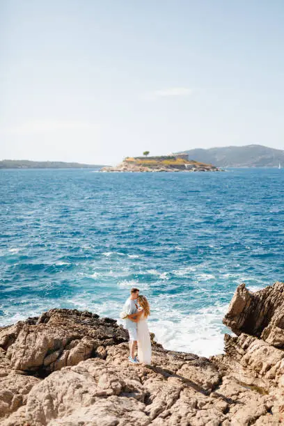 Photo of Man and woman in love stand embracing and holding hands on the rocky seashore, behind them is the Mamula island
