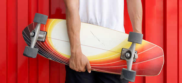 Closeup man's hand holding the surfskate board in hip position in front of red galvanized steel sheet wall.