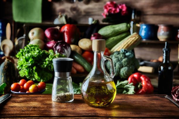 Close-up of olive oil bottle and peppermint on cutting board in rustic kitchen with assorted organic vegetables. Natural lighting: Bell Red Peppers, carrots, lettuce, olive oil, peppermint, potatoes, onions, celery, broccoli, carrots, cucumber, succinic, rucula, chieves, eggplant, corn