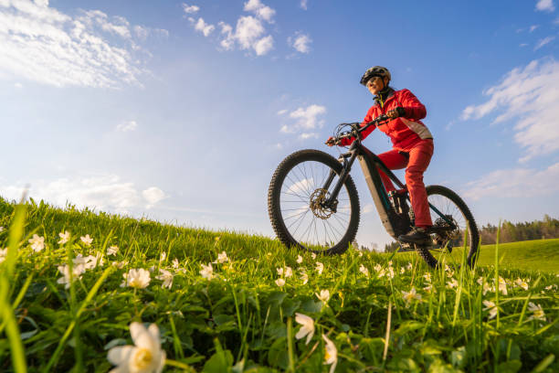 woman on electric Mountain bike with blooming meadow pretty mid age woman riding her electric mountain bike in early springtime in the Allgau mountains near Oberstaufen, in warm  light with blooming spring flowers in the Foreground electric bicycle stock pictures, royalty-free photos & images
