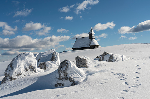 Small wooden chapel covered with snow in Velika planina, Big Pasture Plateau in Slovenia, Europe, in backgrounds mountains against clear blue sky.