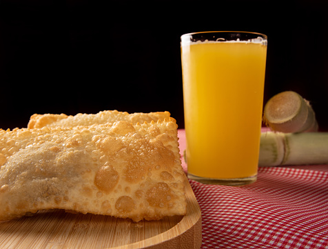 Brazilian fried pastries, a glass of sugarcane juice and canes positioned on a checkered tablecloth, black background, selective focus.