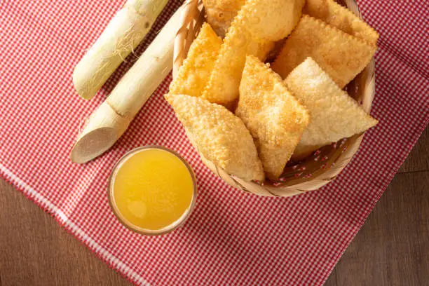 Brazilian fried pastries, a glass of sugarcane juice and canes positioned on a checkered tablecloth, black background, top view.